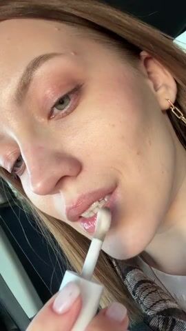 A fair-skinned woman with light brown hair, wearing light make-up, including lip gloss, is shown up close. She sits in a car, wearing a seatbelt. She applies lip gloss with a wand, then smiles at the camera. She has gold chain-link earrings.  Green trees are visible through the car window.