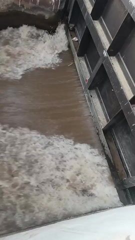The video captures a powerful flow of muddy brown water through a man-made channel, likely in Samundri, Pakistan. The water is highly turbulent, creating significant white foam and rapids as it moves. On the right side of the channel, there is a dark, possibly metallic, structure, which could be part of a gate or barrier, while a white concrete wall forms the left bank. The scene suggests a controlled water flow, possibly from a weir or dam spillway. The video briefly pans upwards at the end, revealing a very bright, overexposed sky or outdoor environment, confirming it's daytime. No people or visible text are present in the video.