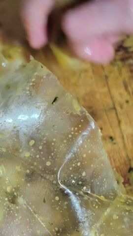 A close-up shot shows a hand cutting open a clear plastic bag containing what appears to be food. The bag is on a wooden surface. The contents of the bag appear to be coated in an oily, yellow substance with small green flecks. The hand uses a silver knife to cut a small slit in the corner of the bag. The lighting is somewhat dim, highlighting the texture of the plastic and the contents within.