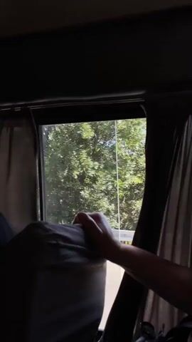 This media file shows a view from inside a vehicle, likely a bus, looking out of a window. The foreground is dominated by a hand resting on the headrest of a seat. The seat itself is covered in a grey fabric. Dark curtains frame the window on both sides.

Through the window, a bright, sunny day is depicted. Lush green trees and foliage fill the middle ground, suggesting a natural or rural setting. In the background, a prominent, rounded mountain or hill rises, its slopes covered in vegetation and appearing dry and arid. A road can be seen in the distance, suggesting movement or travel. The overall scene implies a journey through a scenic, possibly mountainous region with a clear sky and pleasant weather. The focus on the hand and the window view creates a sense of personal perspective and observation of the passing landscape. There is no visible text in the media file. Given the location context of Mukhatgverdi, Georgia, the scene likely depicts the landscape of that region.