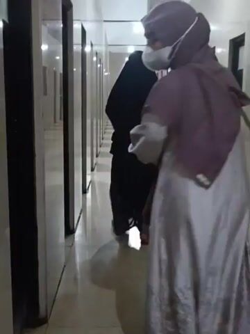 The video shows a hallway with several doors on both sides. A person wearing a black hijab and dress walks down the hallway. Another person, partially visible and facing away from the camera, is in the foreground. This person appears to be wearing a light-colored head covering and a light-colored garment, and is holding a bag. The hallway is lit by overhead lights, and the floor is tiled and appears wet in places. The camera is moving, following the person in the black hijab.