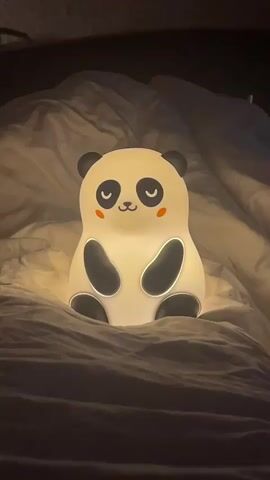 A white silicone panda night light with black markings, a small mouth, closed eyes, and orange cheeks sits on a white bed with wrinkled sheets in a dark environment. The night light emits a warm glow.
