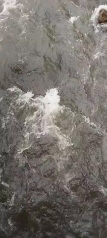 A close-up, overhead shot shows turbulent white water flowing. The water appears to be moving rapidly, with foam and bubbles on the surface. The color of the water is a murky gray-green, with highlights of bright white where the light hits the crests of the waves. The camera angle suggests the viewer is looking down from a high vantage point, perhaps a bridge or a cliff. The overall impression is one of raw, natural power and movement.