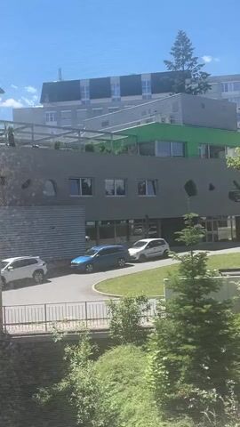 This is a video clip captured from a window, looking out at a modern building complex and its surrounding greenery. The day is bright and sunny, with a clear blue sky visible. The building in the foreground is a multi-story structure with a grey facade, featuring irregular, oval-shaped openings and large windows. A green-roofed section is visible on top of this building, and behind it, a taller building with a white and grey exterior and numerous windows can be seen.

In front of the grey building, a paved area and a grassy lawn are present. Several cars are parked or driving on the paved area, including a white hatchback and a blue sedan. Lush trees and bushes dot the landscape, with a prominent dark green conifer tree in the middle ground, and lighter green deciduous trees further back. The overall impression is of a contemporary urban or semi-urban environment, likely a campus or office park, given the modern architecture and well-maintained grounds. The view suggests it is daytime, possibly late morning or afternoon due to the strong sunlight and shadows. No people are clearly visible, but the presence of cars implies human activity in the vicinity. The video appears to be taken from a slightly elevated position, looking down and across the scene.