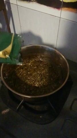 The video captures a dim kitchen scene from an overhead perspective. A metal pot is positioned on a stovetop, containing a quantity of dry, brown, granular material, possibly herbs or spices. A hand, mostly out of frame on the bottom left, holds a green and yellow object, resembling a packet or container. The hand is seen shaking the object, implying that its contents are being added to the pot. The immediate surroundings include a tiled floor with white squares and a colorful mat featuring fruit illustrations (yellow and red) on the left side of the frame. No people or visible text are present. The activity appears to be food preparation.