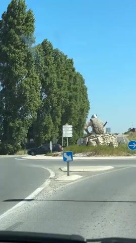 A video, filmed from inside a car, shows a statue of a person sitting on a rock. The statue is large and appears to be made of stone or cement. The person in the statue is wearing a helmet and a fur-like garment. They are holding a stick or spear. There are trees and a blue traffic sign with a white arrow pointing to the right visible in the foreground and background. The car is moving, as indicated by the blur of the surroundings and the slight shift in perspective. The sky is clear and blue, suggesting it is daytime. The car is on a road, likely approaching or passing a roundabout.