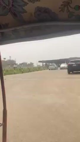 The video shows a view from inside an auto-rickshaw moving along a road.  The rickshaw's patterned fabric roof is visible in the foreground. Outside, the road transitions from paved to unpaved. Signs and a gas station are visible in the background, and a white car is seen driving in the distance.  The sky is hazy.