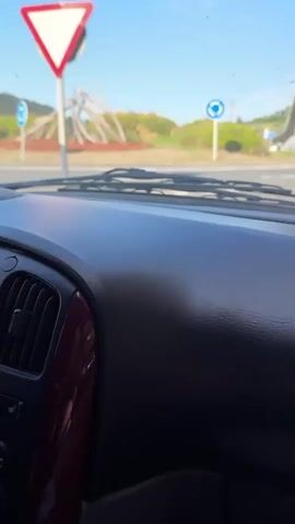 The video provides a first-person perspective from inside a vehicle, looking through the windshield. The dashboard is visible in the foreground, and the windshield wipers are positioned at the bottom of the frame. Outside, a bright blue sky with scattered clouds is visible. In the distance, a blue and white circular traffic sign, indicating a roundabout, is present. The road ahead appears to curve to the right. A concrete overpass or bridge structure is visible on the right side of the frame, casting a shadow on the road. The overall environment suggests an outdoor road setting. There are no people or explicit actions depicted beyond the static view from the car.