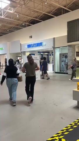The video shows a mall walkway with several people shopping. In the foreground, two young women walk away from the camera, one carrying a shopping bag. Further down the walkway, several other individuals are visible, some walking towards the camera and others away. A store called "WIRELESSWAVE" is prominently displayed with a sign indicating "Under Renovation." The environment appears to be a typical indoor shopping mall with a tiled floor and a high ceiling with exposed pipes and lighting fixtures. There are other storefronts visible in the background, including "CROCS." Yellow and black caution tape is visible in the lower right corner of the frame, suggesting a temporary closure or construction area.