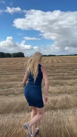 The video shows a woman with blonde hair wearing a denim dress and patterned shoes running through a field of dry, tan-colored stubble under a blue sky with scattered white clouds. The stubble is arranged in long, parallel rows. In the distance, there is a line of trees. The woman is facing away from the camera and appears to be moving towards the horizon.