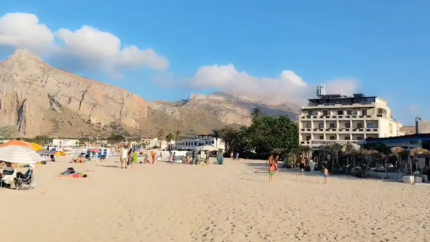 This media file depicts a bustling beach scene in San Vito Lo Capo, Italy, likely during the daytime. The sun is shining brightly, casting a warm glow on the sandy shore, and the sky is a clear blue with a few fluffy white clouds. The atmosphere is one of leisure and enjoyment, as evidenced by the numerous people scattered across the beach.

The subjects of the file are primarily people engaged in various beach activities. Many are sunbathing on towels or in beach chairs, while others are strolling along the shoreline or playing in the sand. Some individuals are relaxing under colorful beach umbrellas, seeking shade from the sun. There are also children present, adding to the lively and family-friendly atmosphere.

Objects visible include beach umbrellas, towels, beach chairs, and some buildings in the background, suggesting a developed coastal resort area. One prominent building is a multi-story hotel with visible balconies, and next to it, what appears to be a restaurant with an outdoor seating area under a thatched roof.

The setting is a wide, sandy beach meeting the calm, blue waters of the sea. In the background, a dramatic, imposing mountain looms over the scene, its rocky slopes catching the sunlight. This distinctive mountain, likely Monte Monaco, is a key geographical feature of San Vito Lo Capo and serves as a significant location cue. The presence of palm trees also contributes to the Mediterranean ambiance.

The overall mood conveyed is relaxed and happy. The people appear to be enjoying a summer day at the beach, seeking recreation and relaxation. The vibrant colors of the umbrellas and the blue of the sky and sea create a cheerful and inviting scene.

There is visible text on the hotel building that reads "RISTORANTE" in black lettering.