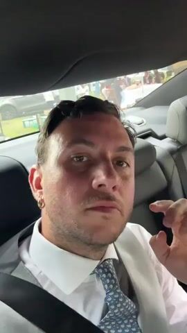 A man with dark hair and a beard, wearing a white shirt and a light grey vest with a blue patterned tie, is sitting in the backseat of a car. He has a large hoop earring in his left ear. The man is looking at the camera and touches his forehead. Another man, with short hair and wearing a white shirt and a tan vest with an orange tie, is sitting next to him. The second man is smiling at the camera and has his hands on the steering wheel. The interior of the car is visible, including the grey leather seats and the black headliner. Outside the car window, trees and buildings can be seen. The lighting suggests it is daytime.