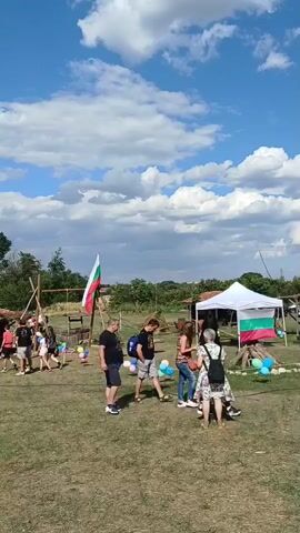 The video shows an outdoor gathering with people milling around a grassy field. A Bulgarian flag is prominently displayed on a wooden pole, and another smaller Bulgarian flag is visible on a white tent. There are several people walking and standing, some interacting with each other, while others appear to be observing. The environment seems to be a rural setting with trees and buildings in the background. The sky is blue with scattered clouds, suggesting it is daytime. The overall atmosphere appears to be casual and festive, with balloons scattered around the area. The people are dressed in casual clothing.