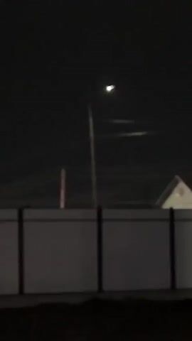 This media file captures a nighttime scene in Qoyankus, Kazakhstan. The primary focus is a tall street lamp with its light illuminating the dark sky. Below the lamp, a long, grey fence with dark vertical posts stretches across the frame. Behind the fence, the outline of a building, likely a house with a triangular roof, is faintly visible. The sky is dark, suggesting it is nighttime. There are no people or animals visible in the frame. The overall mood is quiet and still, typical of a deserted street at night. There is no visible text.