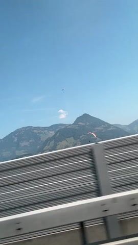 The video shows a view from a vehicle traveling on a road. In the foreground, there is a modern, gray sound barrier with horizontal lines. Beyond the barrier, a clear blue sky is visible with a few wispy clouds. In the middle ground, a mountain range with green slopes and trees is present. Two paragliders are visible in the sky; one is higher up and appears smaller, while the other, larger and orange and white, is closer to the mountains. The trees on the slopes are a mix of green and some with yellow and orange autumn colors, indicating the season. The video captures a scenic view of mountains and the activity of paragliding.