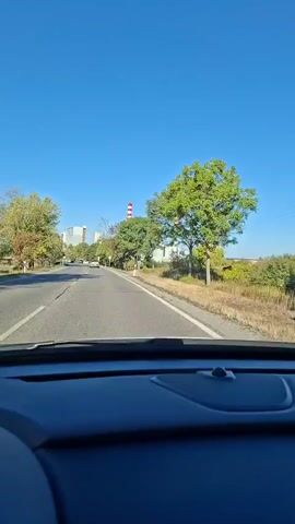 A dashcam view shows a car driving down a paved road on a clear, sunny day. The road has a white line dividing the lanes and a white line along the shoulder. Trees with green and yellow leaves line both sides of the road. In the distance, there are several buildings and a tall, red and white tower. There are a few other cars on the road.