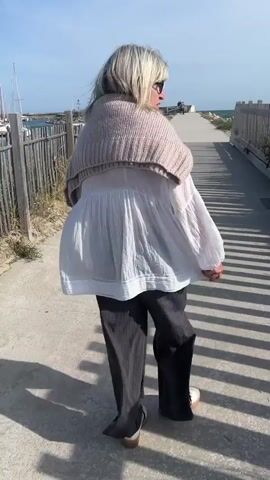 A woman with gray hair, wearing a beige knit wrap, a white blouse, and dark pants, walks away from the camera on a concrete path towards the ocean. The path is lined with wooden fences on both sides, casting long shadows.  The ocean and sky are visible in the distance.  On the left side of the path, behind the fence, is a marina with boats.