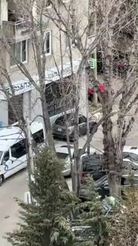 From an elevated position, the video provides a view of a parking area and adjacent street framed by bare trees in the foreground. Cars of various makes and models are parked in the lot. A building is partly visible on the left with signage in a non-English alphabet. A red object, possibly a piece of fabric, is caught in the branches of one of the trees. As the camera pans slightly to the right, a narrow lane lined with garages or sheds becomes visible. The scene appears to be in an urban or suburban area, on a somewhat overcast day.