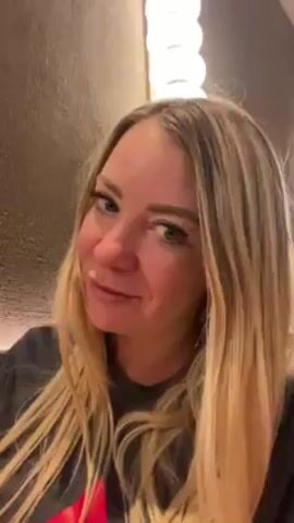 The video shows a woman with blonde hair, who appears to be in her 30s, drinking red wine from a large glass. The lighting is warm, possibly inside a restaurant, with a textured wall behind her and a string of lights visible at the top of the frame. She looks directly at the camera before and after taking a sip of the wine. The setting seems casual and intimate.