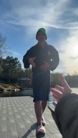 A man stands on a wooden dock next to a body of water under a bright, sunny sky. He wears a dark blue bathrobe with a patch resembling the Russian double-headed eagle coat of arms, and a green knit hat. He extends his right hand towards the camera, smiling broadly. The camera, held by another person, moves in, and their hand meets the man's hand for a handshake. The sun creates a strong lens flare behind the man during the handshake. Following the handshake, the man's expression subtly shifts, suggesting a reaction to the cold or the sensation of the touch. The background features a large body of water, lined with trees and some distant buildings, under a clear blue sky with a few clouds. The scene is set outdoors, likely by a lake or river in Shchelkovo, Russia, on a bright day.