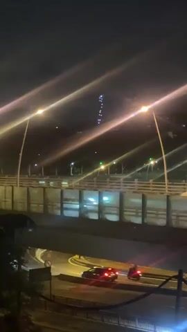 The video captures a nighttime scene of a multi-level road system, likely in Saint-Cloud, France. In the foreground, below a concrete overpass, a dark sedan with red taillights and a motorcycle with a single red taillight are seen moving from left to right on a lower road. Above them, on the bridge structure, a light-colored car, possibly a white sedan, is visible moving to the right, its form slightly blurred by motion.

The scene is illuminated by numerous streetlights positioned along the bridge, creating prominent lens flares that stretch diagonally across the frame. In the distant background, a tall, slender structure, possibly a tower or antenna, is visible, adorned with multiple blinking or sequentially lighting blue and white lights. The sky is dark, indicating it is nighttime. There are no other notable weather conditions. No text is visible in the video.