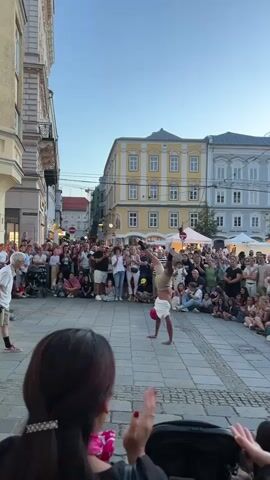 The video shows a street performance in a European city square. A breakdancer is performing in the center of the square, surrounded by a large crowd of spectators. The dancer is wearing a white t-shirt and khaki shorts and is executing a handstand. To the left of the dancer, another performer, an elderly man wearing a white shirt and shorts, stands watching. The buildings surrounding the square are old and ornate, with yellow and gray facades. The sky is clear and blue, indicating it is daytime. The crowd is diverse, with people of all ages and backgrounds. Some people are sitting on the ground, while others are standing. The atmosphere appears festive and engaging.
