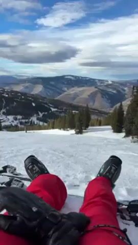 A person is sitting on a snowy mountain slope wearing red ski pants and ski boots. They are positioned with their legs outstretched towards the camera. The person is also wearing ski gloves, one of which is visible in the foreground. Beyond the person, the snow-covered slope stretches down towards a valley with coniferous trees and distant mountains under a partly cloudy sky. Another person, wearing a white and black ski outfit, walks past the first person and then stops at the side of the slope.