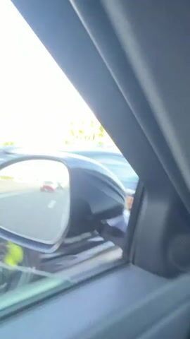 The video shows a view from inside a car, looking out the side mirror. The side mirror reflects a convoy of cars driving on a highway. The cars are traveling in the same direction as the camera car. The landscape outside the car consists of trees and a clear blue sky. The sun appears to be setting, casting a warm glow on the scene. One car in the reflection, a red sports car, is prominent. Another car, a black sports car, is also visible in the reflection, in front of the red car. The road is paved and appears to be in good condition. There are other cars visible in the distance in the reflection. The camera car is also moving, as indicated by the blur of the scenery outside the window.