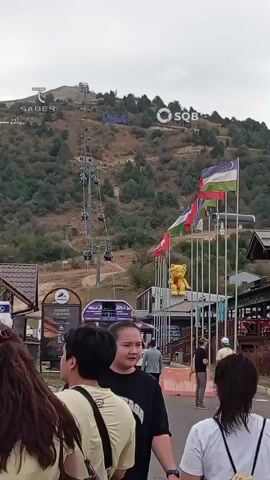 The video shows a group of people standing in what appears to be a tourist area at the base of a mountain with a ski lift. In the background, the ski lift is visible ascending the mountainside. Several flags are displayed on poles, including what appears to be the flag of Uzbekistan. A large yellow teddy bear statue is also present. People in the foreground are facing away from the camera, and some are holding cell phones. The ground is paved, and there are buildings and signs around the area. The overall scene suggests a recreational or resort setting.