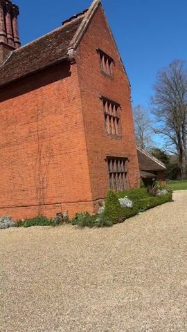 The video shows a multi-story brick building on a sunny day. One section of the building is almost entirely covered in ivy. There are several windows with panes and a roof tiled in brown. A chimney rises above the roofline. The building features a large arched entranceway. In front of the building is a gravel driveway where a black convertible car is parked. The sky is blue and clear.