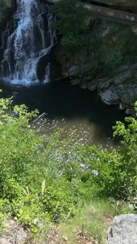 This media file captures a scenic view of a waterfall cascading into a dark pool of water. The scene is set within a rocky, natural environment with lush green foliage framing the foreground and surrounding the waterfall. The water from the falls creates ripples and a shimmering effect on the surface of the pool, suggesting sunlight glinting off the water.

The waterfall itself appears to be a multi-tiered cascade, with water flowing over rocks and creating a dynamic visual. To the left of the main waterfall, there is a man-made structure resembling a dam or retaining wall with several levels from which water is also flowing down, suggesting a controlled or enhanced water flow. The presence of concrete structures and what appears to be a walkway or path along the top of the rock face indicate that this location is accessible and possibly a tourist attraction.

The time of day appears to be daytime, with ample sunlight illuminating the scene. The weather seems pleasant, with clear skies implied by the bright lighting. The location cues, such as the rocky terrain and the type of vegetation, along with the mention of Užice, Serbia, place the scene in a natural, possibly mountainous region.

No people are visible in the media file. The primary subjects are the natural elements of the waterfall and the surrounding landscape, along with the man-made structures that interact with the water flow. The overall emotion evoked is one of natural beauty and tranquility, with a touch of human intervention evident in the infrastructure. No visible text is present in the media file.