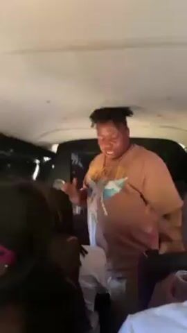 Inside a vehicle, possibly a van, a man wearing a tan graphic t-shirt with an image of an angel, khaki shorts, and dreadlocks stands up and dances. He moves his arms and body to the rhythm of the music, facing the camera. Other passengers are visible in the background, including a woman with a pink headband and a man with a white and green baseball cap. The interior of the vehicle is white, and the back of the seats are dark. The scene appears to be lively and energetic.