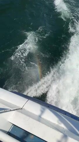 The video shows the view from a boat moving through the water. The water is dark green and choppy, with white foamy waves created by the boat's movement. A small rainbow appears in the spray of water near the boat's edge. The camera is positioned on the boat, looking down at the water. Part of the boat's white railing is visible at the bottom of the frame.