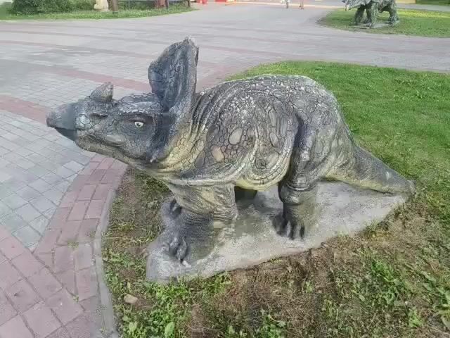 A statue of a triceratops dinosaur is shown in this video. The statue is gray and yellow, with green eyes. It is standing on a rock in a grassy area next to a brick pathway. The statue is facing to the left.