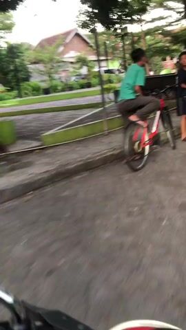 The video shows two young males with bicycles in an outdoor setting, possibly a park or residential area. The first male is riding a red bicycle, wearing a teal shirt and brown pants, and appears to be moving away from the camera. The second male is standing to the right, wearing a dark shirt and shorts, and is also near a bicycle. The ground is paved with an asphalt-like material, and there is a green-painted barrier in the foreground. Trees and some structures are visible in the background, suggesting a daytime scene with natural light.