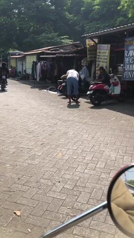 The video shows a street scene with several market stalls lining the right side of the frame. The stalls are covered by awnings and display various goods, including clothing and what appear to be food items. Several motorcycles are parked or being ridden in the street. In the foreground, a person wearing a white t-shirt and blue shorts is standing. Further down the street, other individuals are visible near the stalls and motorcycles. The ground is paved with interlocking bricks, and trees are visible in the background, suggesting an outdoor environment. The lighting indicates it is daytime, likely sunny.