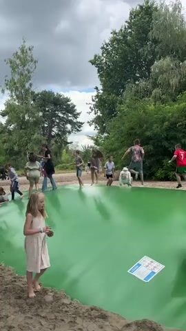 The video shows a group of people, mostly children, playing on a large, inflatable green trampoline surrounded by sand. The trampoline is set outdoors in a wooded area with trees and foliage visible in the background. The sky is cloudy. People are jumping, running, and interacting on the trampoline. Some adults are observing from the edges. There are also a few pairs of shoes scattered on the sand near the trampoline.