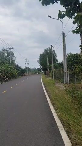 The video is a first-person perspective recording of a road with a cloudy sky. A paved road stretches into the distance with a white line on the right edge. On the left side of the road, there are trees, utility poles with wires, and a motorcycle. On the right side, there is a fence, more trees, and grass. A large white tanker truck with blue and red accents passes by, moving away from the camera. The truck has writing on its side, including the words "Cherry Valley" and a license plate that reads "60C-736.06".