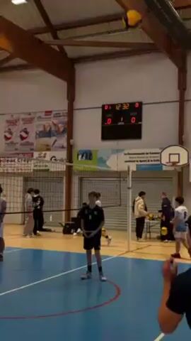 Several volleyball players are engaged in a practice session on an indoor court with a blue surface and a net stretched across the center. The volleyball is yellow and blue, and it is in the air above a player getting ready to serve. The score on a digital scoreboard indicates the match is in progress. The walls of the gymnasium are white with posters and advertisements displayed. The players are wearing different colored uniforms.