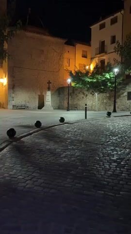 The video captures an empty, cobblestone plaza at night. Streetlights illuminate the scene, casting shadows on the ground. A stone monument with a cross stands in the background, and buildings with illuminated windows line the plaza. A tree and a motorcycle are also visible. The atmosphere is quiet and still.