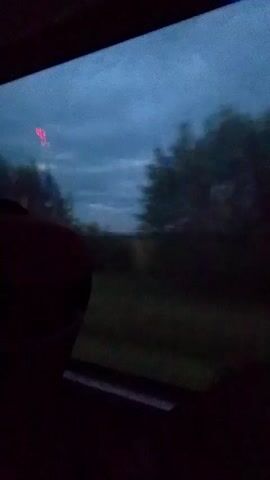 The video shows a view from a moving vehicle, likely a bus or train, looking out a window. The sky is overcast with dark clouds, suggesting twilight or stormy weather. Green foliage and trees are visible outside, blurring due to motion. A dark interior of the vehicle is visible in the foreground, with a red headrest or seatback on the left. There are some indistinct red lights visible in the distance, possibly from streetlights or vehicles.
