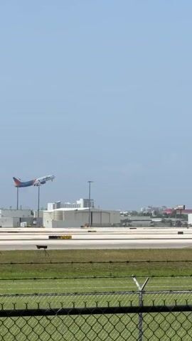 An airplane with a blue, orange, and white livery is ascending against a clear blue sky. The aircraft appears to be a commercial jetliner. Below, an airport tarmac is visible, with several other parked aircraft, including a white private jet and a blue and white commercial jet. Airport buildings and hangars are also present in the background. In the foreground, a chain-link fence and a grassy area are visible. The scene suggests an active airport environment on a sunny day.
