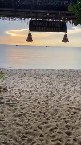The video shows a beach scene, framed by the sign for Wangsai Resort & Restaurant and a thatched roof. The beach is sandy, with a person lying on a towel in the distance. The water is calm, reflecting the colors of the sunset, and two boats are visible on the horizon. The sky is a mix of clouds and sunlight.