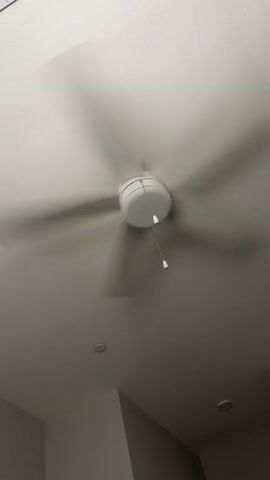 The video shows a close-up, low-angle shot of a white ceiling fan with five blades. The fan is in motion, with the blades appearing as a blur due to their speed. A white cord with a white pull chain hangs from the center of the fan. The ceiling is white and textured. Two circular recessed lights are visible on the ceiling below the fan. The walls in the lower part of the frame are painted a light color and appear to be angled. The lighting is dim and appears to be artificial.