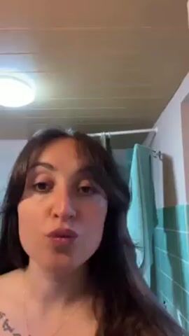 The video shows a person with shoulder-length brown hair and bangs, wearing a white-and-red bracelet, applying eyebrow makeup. The person is holding a small, thin brush with a black handle and light-colored bristles and using it to apply makeup to their left eyebrow. The background is a bathroom with a white ceiling, a round light fixture, and green tiles on the wall to the right. A towel rack with a towel hanging is visible on the right side of the frame. The person is looking directly at the camera.