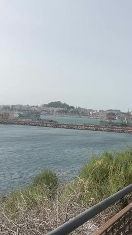 This video shows a panorama of a coastal city from a slightly elevated viewpoint. In the foreground, there is a metal railing and some dry, brush-like vegetation. The middle ground consists of a large body of water, possibly a harbor or bay, with ripples on its surface. Several ships, including what appear to be naval vessels, are docked or anchored in the water. Beyond the water, the cityscape is visible, with buildings clustered along the shore and extending inland. In the background, hills and mountains form a natural backdrop to the city. The sky is bright and hazy, suggesting a sunny day, possibly with some atmospheric dust or pollution. The camera pans slowly across the scene, offering a wide view of the harbor and the surrounding landscape. There are no people or animals visible in the video. The overall impression is one of a bustling port city on a clear, warm day.