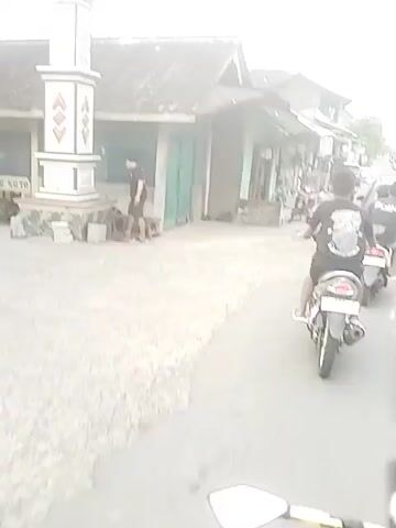 The video shows a person riding a motorcycle. The person passes by a small blue stand with a sign on it. The motorcycle continues down a narrow street with buildings on either side. There are other motorcycles and a car parked along the street.  The sky is overcast.