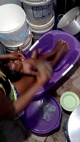 In the video, a person is bathing a baby. The baby is lying in a purple plastic bathtub. The person is using a sponge to wash the baby. There is a bowl of water next to the tub. There are also several white buckets stacked up in the background. The person is wearing a colorful top. The baby appears to be dark-skinned. The scene appears to be indoors.