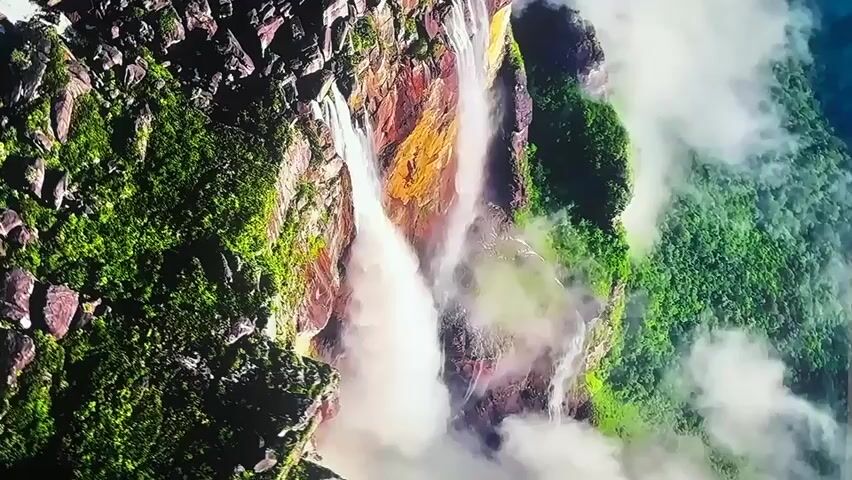 This is a video of Angel Falls in Venezuela, taken from a drone or helicopter. The camera is looking down on the falls, which are a series of cascades that drop over a cliff face. The water is white and foamy, and there is a lot of mist and spray. The surrounding vegetation is lush and green, and the cliffs are a reddish-brown color. The video is oriented vertically, so the falls appear to be running sideways.