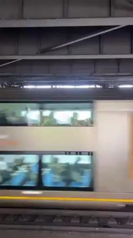 The video shows a train passing by at a train station. The train is a double-decker train with blue seats visible inside. After the train passes, a view of the Sydney Harbour Bridge becomes visible through the opening of the train station. The bridge is a large steel arch bridge, and behind it is the skyline of Sydney. The day appears overcast.